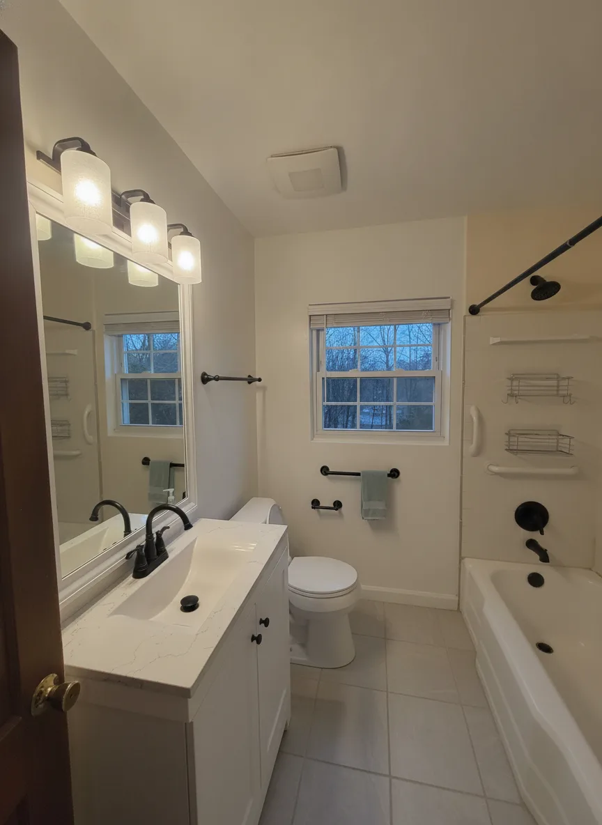 The same bathroom with white walls, large-format gray floor tile, matte black fixtures and a white shaker vanity with quartz top